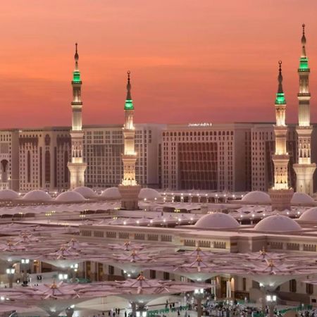 Intercontinental Dar Al Iman Madinah By Ihg Hotell Medina Exteriör bild The photo depicts a scenic view of a mosque complex, likely during sunset, with several minarets illuminated and showcasing green lights. The architecture features domed roofs, and the setting includes a backdrop of buildings, all bathed in a warm, o