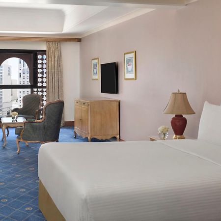 Intercontinental Dar Al Iman Madinah By Ihg Hotell Medina Exteriör bild The photo shows a well-furnished hotel room. It features a large bed with a white duvet, two bedside lamps, and decorative paintings on the walls. There is a comfortable seating area with two chairs and a small table. A wooden dresser is positioned a