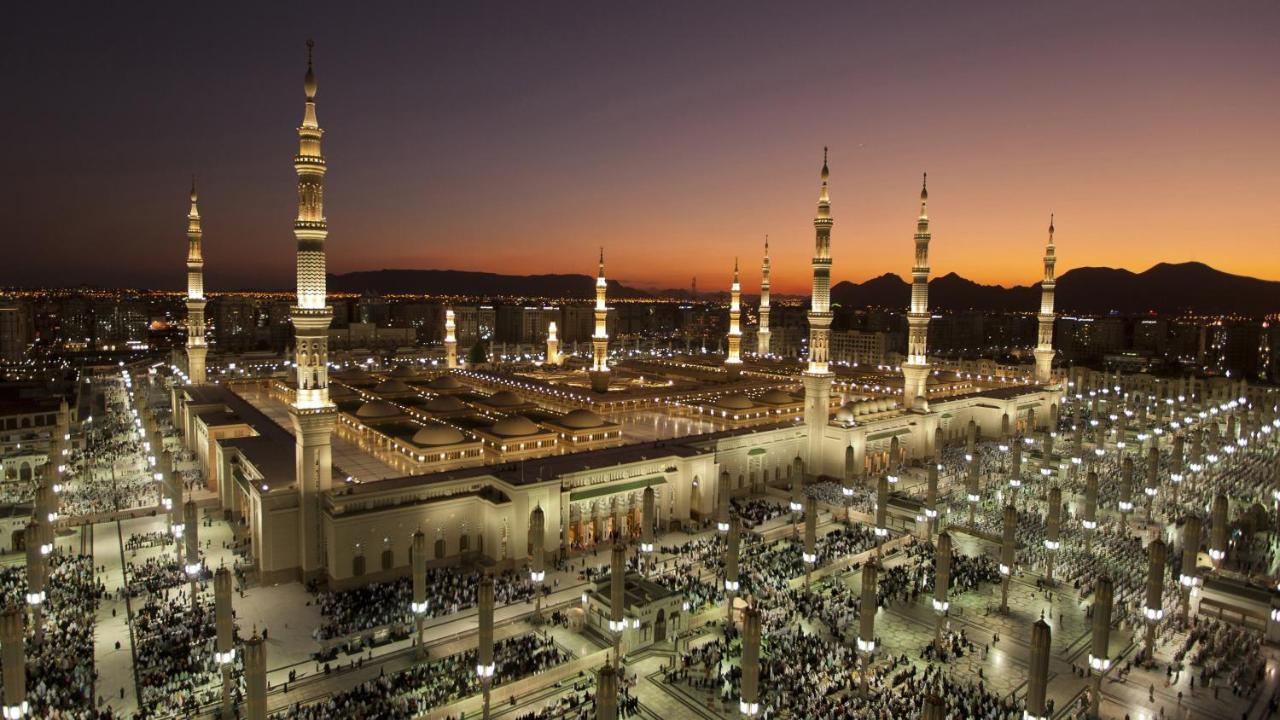 Intercontinental Dar Al Iman Madinah By Ihg Hotell Medina Exteriör bild The photo depicts an aerial view of a large mosque complex during sunset. The mosque features multiple tall minarets, which are intricately designed and illuminated, giving the scene a stunning appearance as the sky transitions from daylight to dusk.