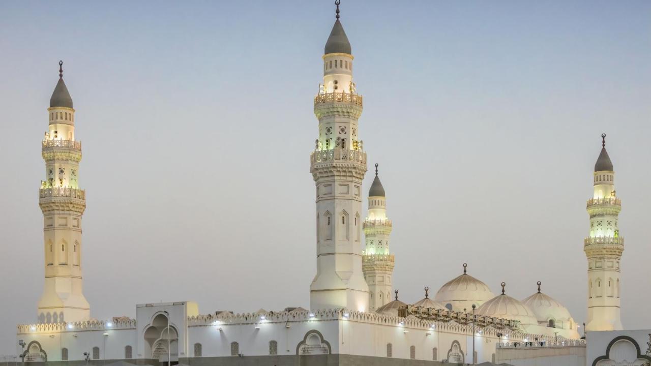 Intercontinental Dar Al Iman Madinah By Ihg Hotell Medina Exteriör bild The image shows a mosque with distinct architectural features, including tall minarets and domes. The minarets are highlighted, likely lit up in the evening, showcasing intricate design elements typical of Islamic architecture. The overall scene conv