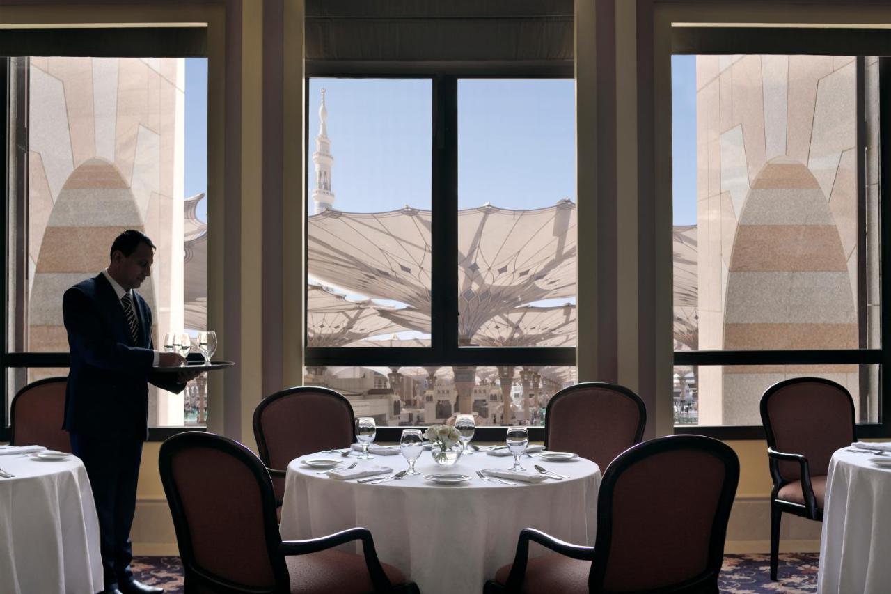 Intercontinental Dar Al Iman Madinah By Ihg Hotell Medina Exteriör bild The photo shows a dining area with elegant decor. A waiter is standing to the side, holding a tray. The tables are neatly set with white tablecloths and fine dining arrangements. Large windows provide a view of an outdoor area featuring architectural