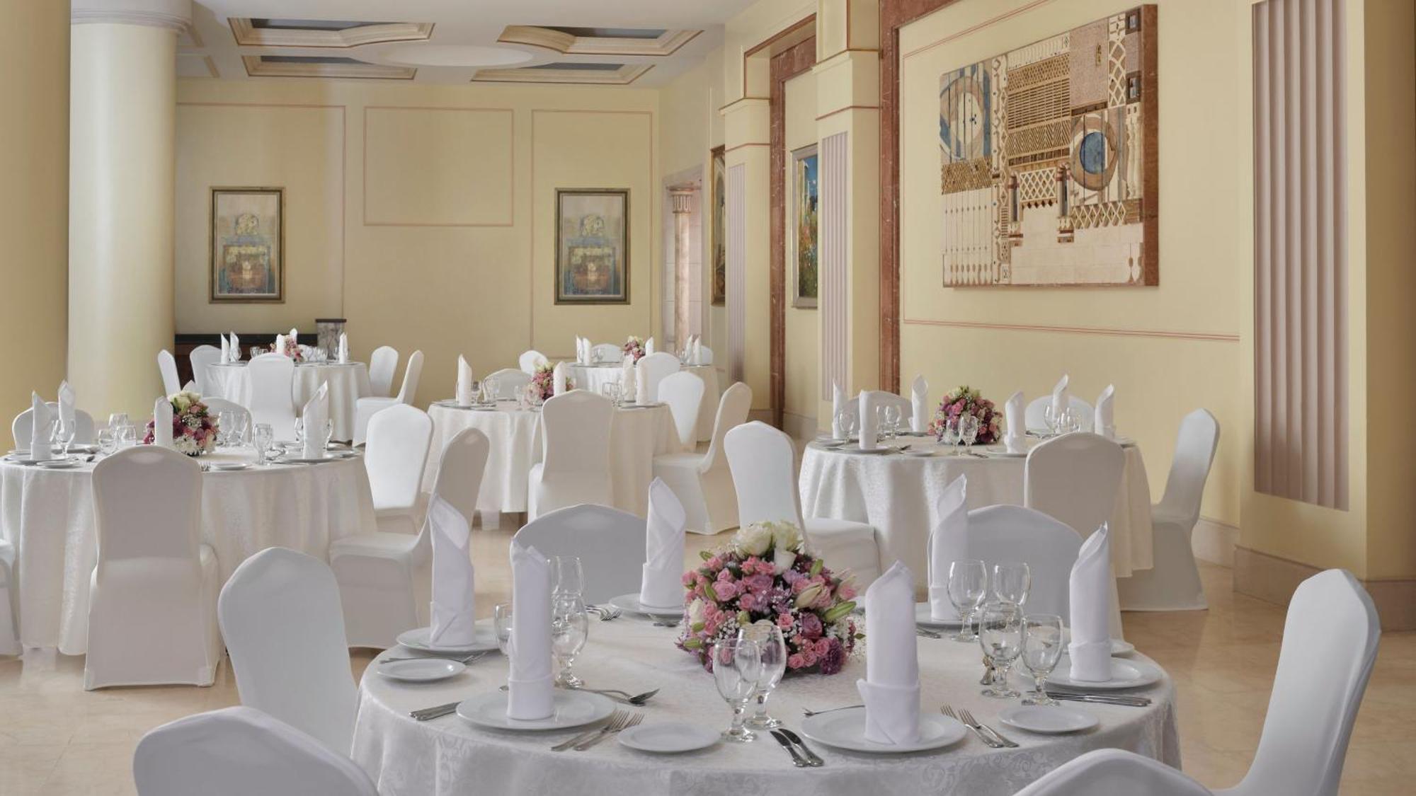 Intercontinental Dar Al Iman Madinah By Ihg Hotell Medina Exteriör bild The photo shows an elegantly arranged dining area, likely set up for a formal event or reception. There are several round tables covered with white tablecloths, each adorned with floral centerpieces featuring pink flowers. The tables are set with pla