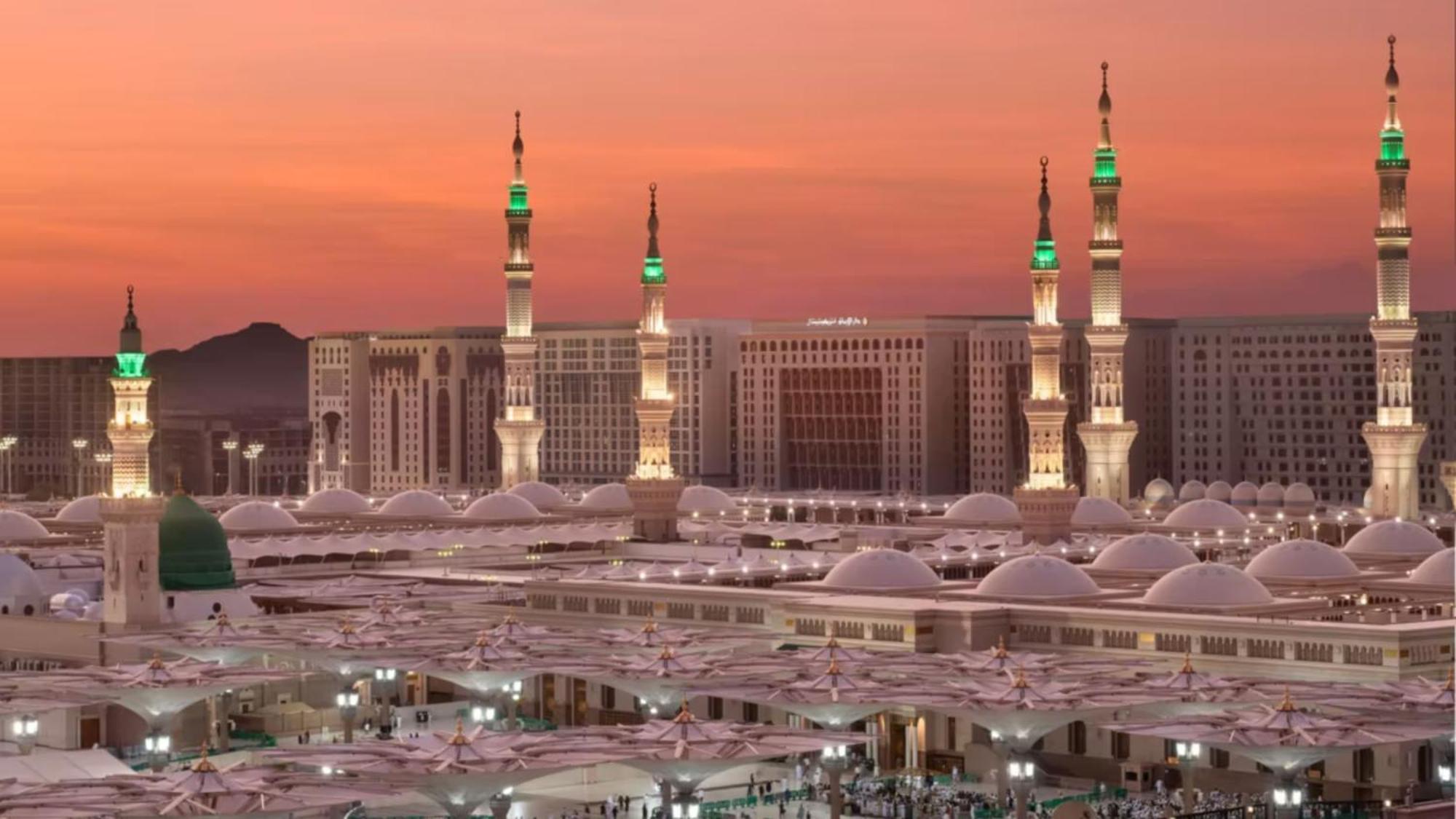Intercontinental Dar Al Iman Madinah By Ihg Hotell Medina Exteriör bild The photo depicts a scenic view of a mosque complex, likely during sunset, with several minarets illuminated and showcasing green lights. The architecture features domed roofs, and the setting includes a backdrop of buildings, all bathed in a warm, o