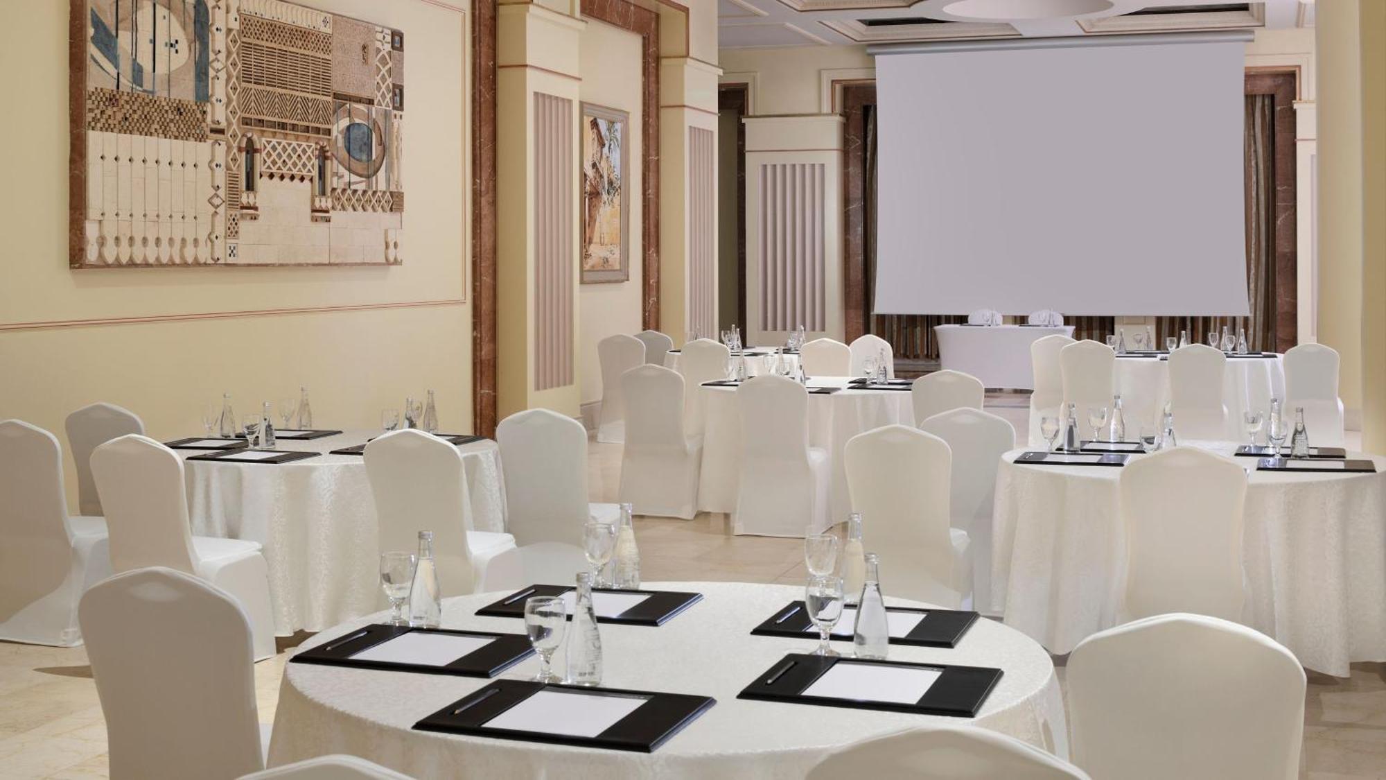 Intercontinental Dar Al Iman Madinah By Ihg Hotell Medina Exteriör bild The photo depicts an elegantly arranged event space. There are several round tables covered with white tablecloths and surrounded by white chairs. Each table is set with a black folder or menu and a glass of water. The walls are adorned with artworks