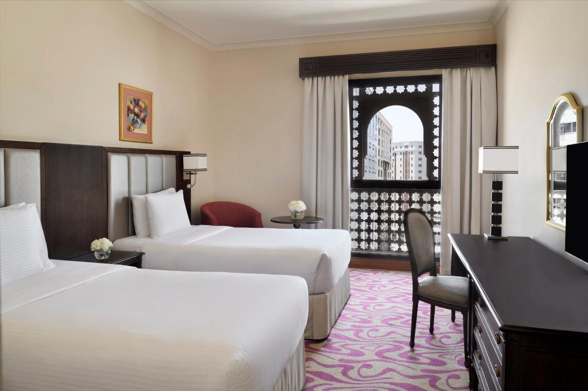 Intercontinental Dar Al Iman Madinah By Ihg Hotell Medina Exteriör bild The photo shows a hotel room featuring two single beds with white bedding. The room has a light, neutral color scheme and is decorated with minimal furnishings. There's a small seating area with a red chair and a round table, as well as a desk with a
