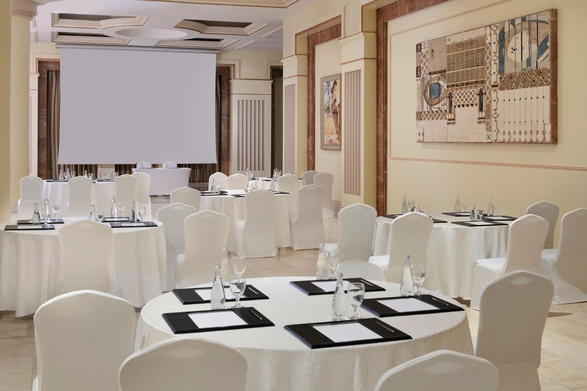 Intercontinental Dar Al Iman Madinah By Ihg Hotell Medina Exteriör bild The photo shows a well-decorated event space or meeting room. It features several round tables covered with white tablecloths and surrounded by white chairs. Each table has black placemats and glass water bottles, suggesting a formal setup for a gath