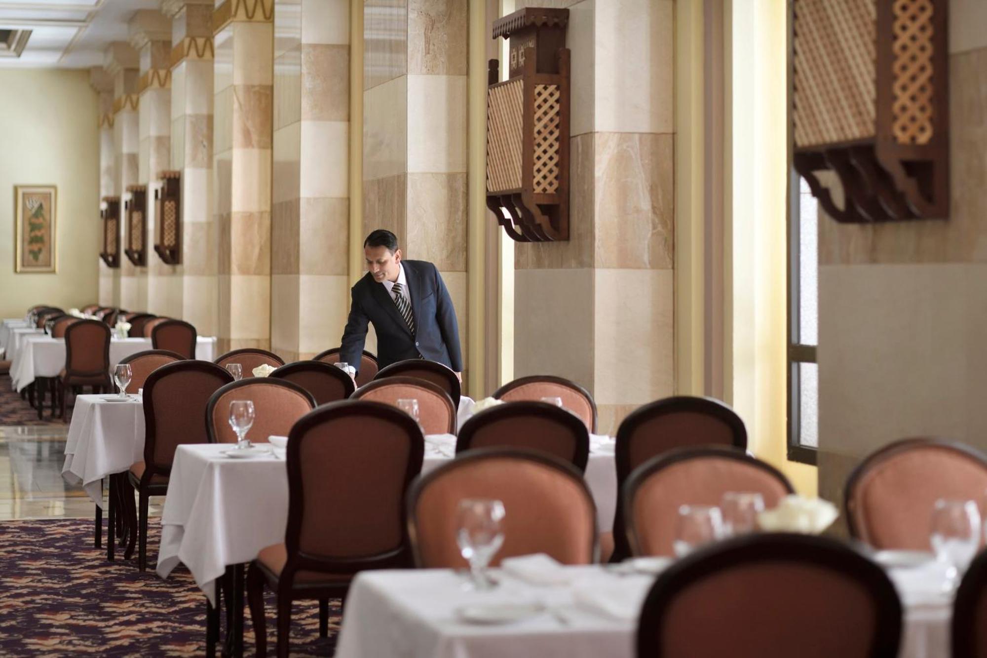 Intercontinental Dar Al Iman Madinah By Ihg Hotell Medina Exteriör bild The photo depicts a dining area in an elegant restaurant. There are several tables set with white tablecloths, each accompanied by chairs. A waiter, dressed in a formal suit, is seen arranging or tending to one of the tables. The restaurant features 