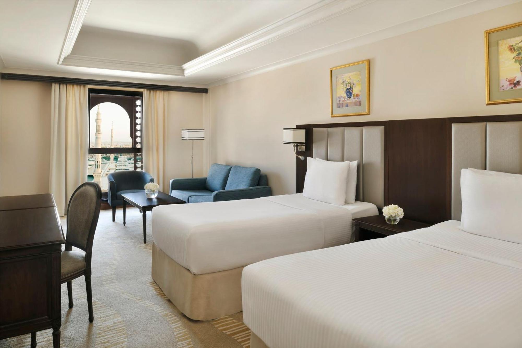Intercontinental Dar Al Iman Madinah By Ihg Hotell Medina Exteriör bild The photo shows a well-appointed hotel room featuring two twin beds with white bedding. There is a small desk with a chair, a comfortable blue sofa, and a coffee table in the sitting area. The room has a warm, neutral color scheme, accented by two pa