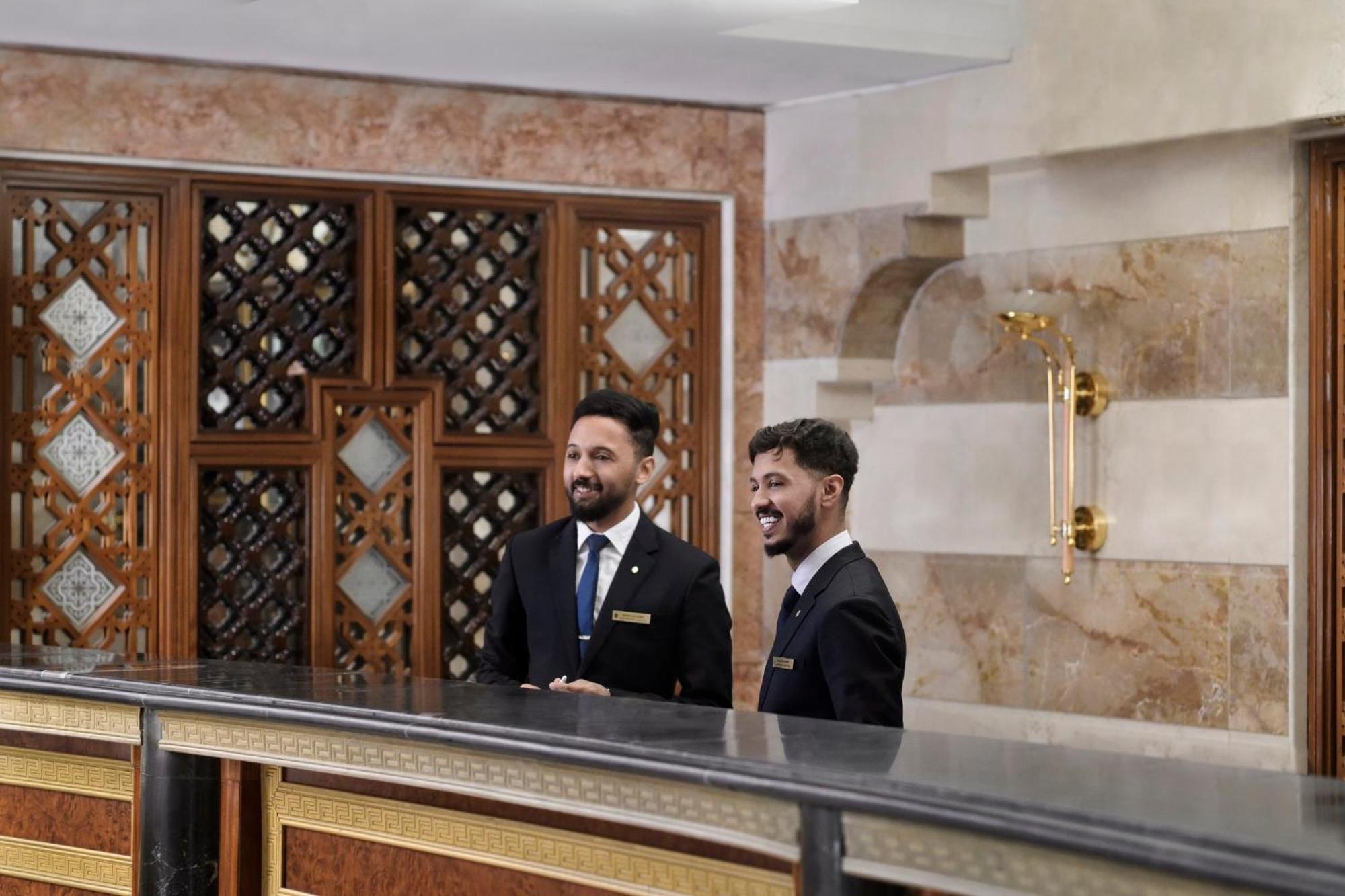 Intercontinental Dar Al Iman Madinah By Ihg Hotell Medina Exteriör bild The photo shows two men standing behind a reception desk in an elegant setting, likely a hotel or similar establishment. They are both dressed in formal attire, wearing suits and ties, and appear to be smiling or engaging in conversation. The backgro