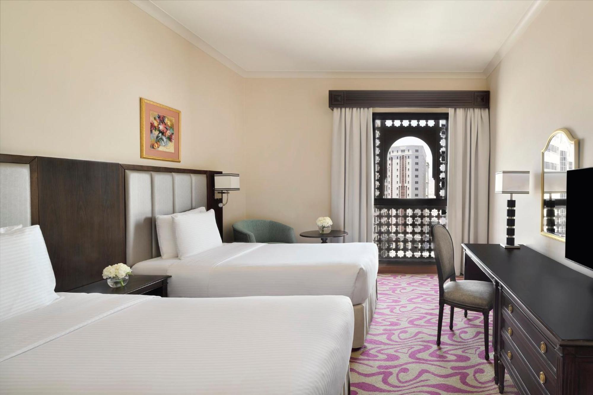 Intercontinental Dar Al Iman Madinah By Ihg Hotell Medina Exteriör bild The photo shows a hotel room featuring two twin beds with white bedding. There is a decorative piece of art on the wall above one of the beds. A small round table with a flower arrangement is placed next to the beds. A desk and chair are situated aga