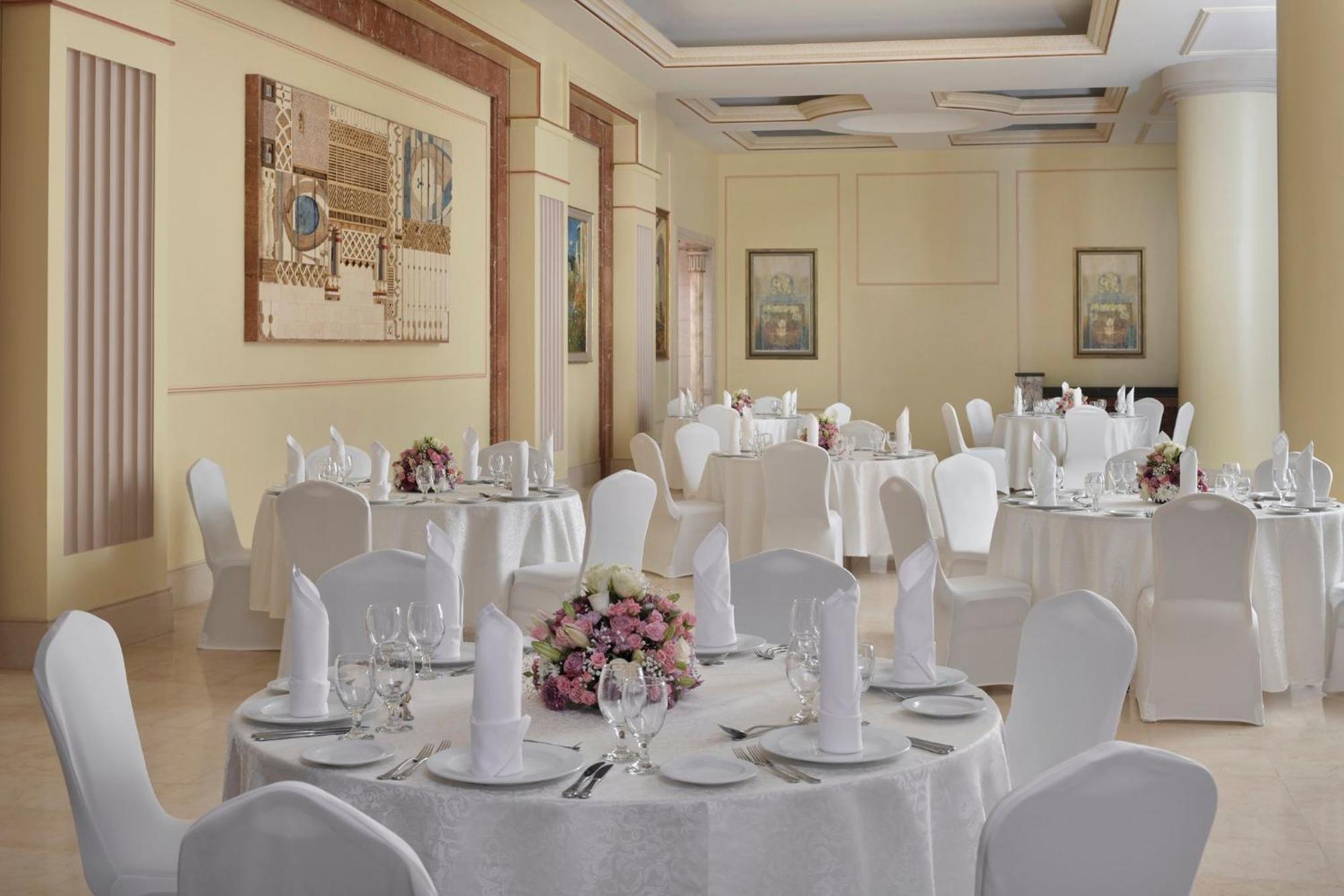 Intercontinental Dar Al Iman Madinah By Ihg Hotell Medina Exteriör bild The photo shows an elegantly arranged dining or function room. There are round tables covered with white tablecloths, each set with fine dining ware including glassware, utensils, and neatly folded white napkins. Some tables have floral centerpieces 