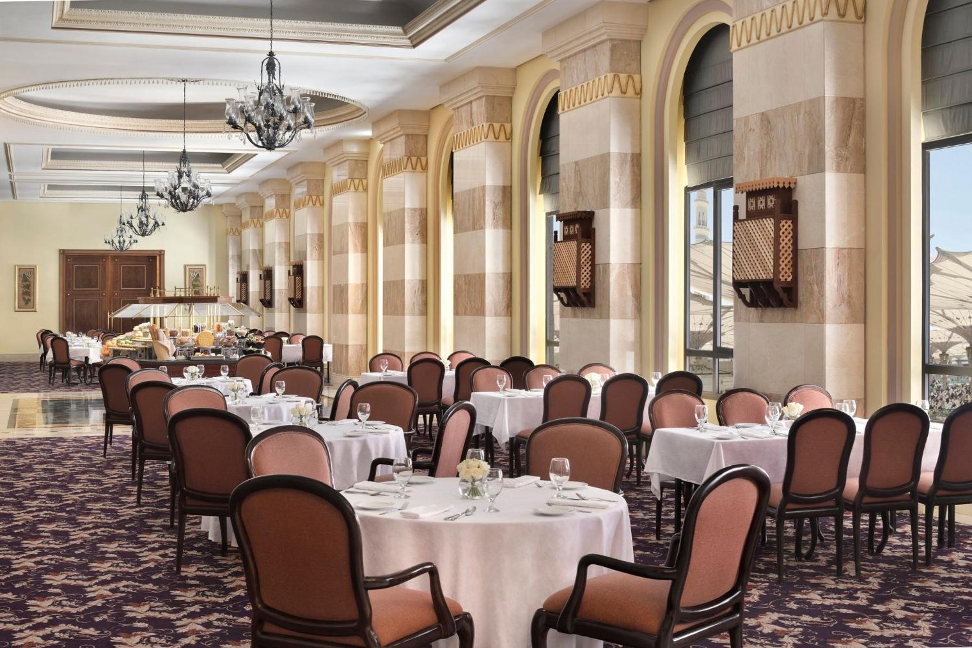 Intercontinental Dar Al Iman Madinah By Ihg Hotell Medina Exteriör bild The photo depicts an elegant dining room setting. It features several round tables covered with white tablecloths, each set with dishes and glassware. The room has a sophisticated ambiance, characterized by high ceilings adorned with chandeliers and 