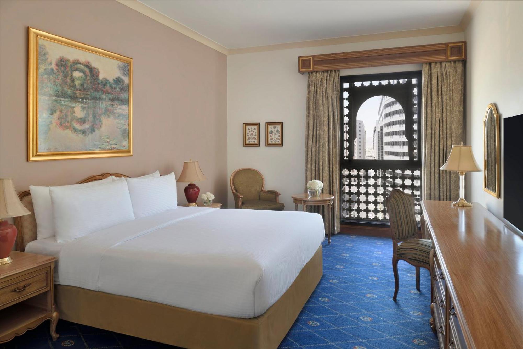 Intercontinental Dar Al Iman Madinah By Ihg Hotell Medina Exteriör bild The photo shows a hotel room that features a large, neatly made bed with white linens. There is a framed painting on the wall above the bed. To the side, a small seating area includes a chair and a side table. Opposite the bed, there is a wooden desk