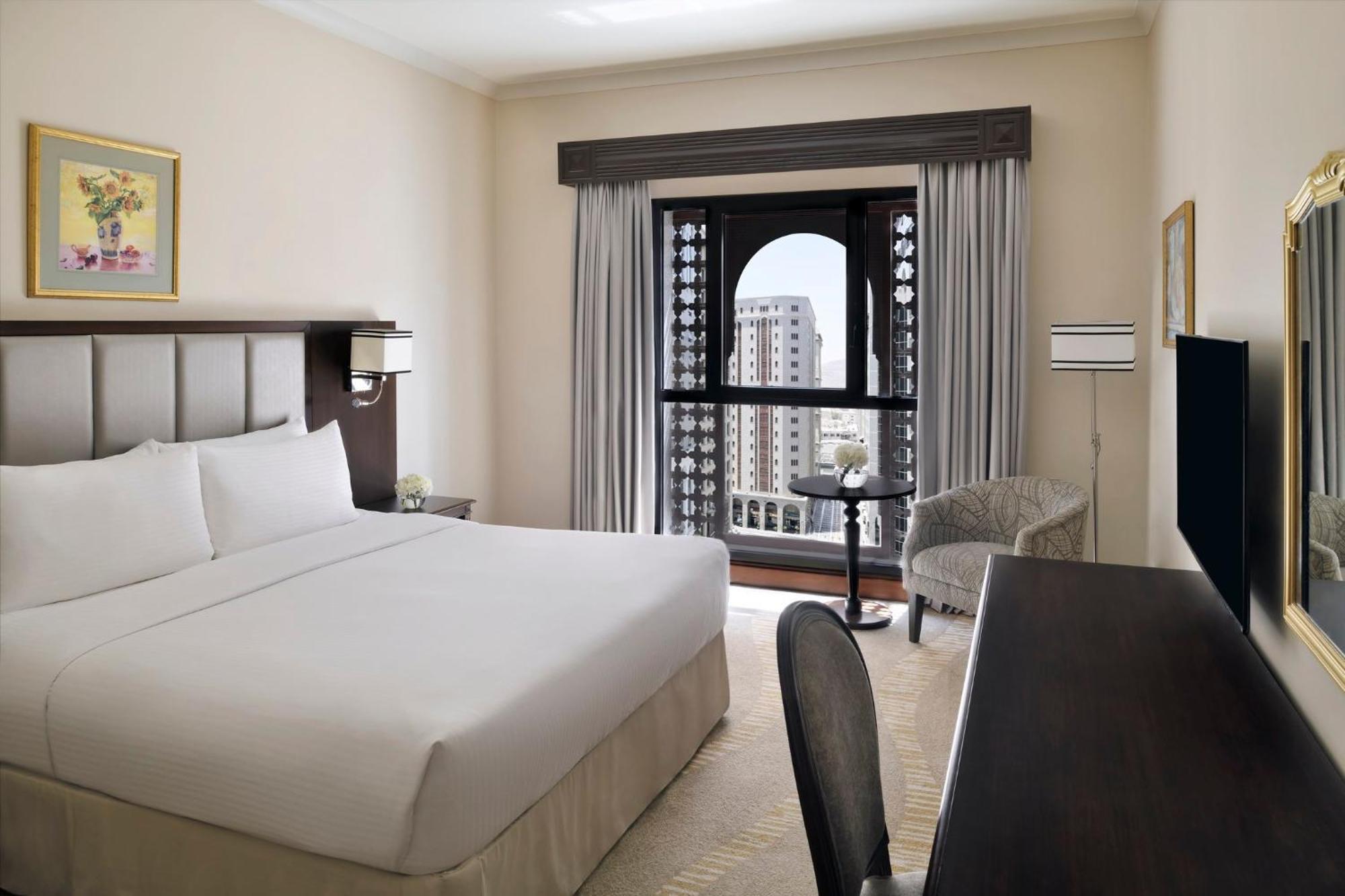 Intercontinental Dar Al Iman Madinah By Ihg Hotell Medina Exteriör bild The photo shows a hotel room with a modern and minimalistic design. It features a large bed with white linens, a side table, and a comfortable chair near a window. The window has a view of a cityscape, and elegant curtains frame the window. There is 