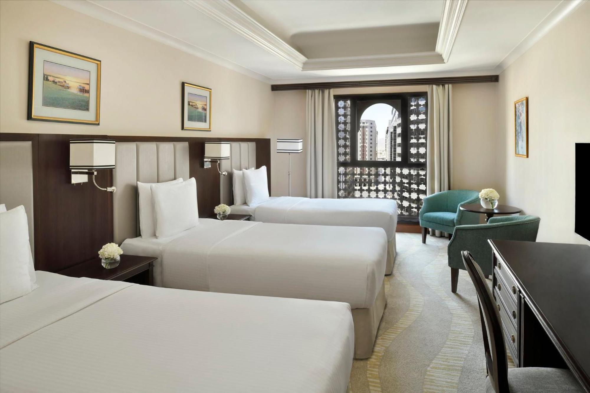 Intercontinental Dar Al Iman Madinah By Ihg Hotell Medina Exteriör bild The photo shows a hotel room with three single beds, dressed in white linens. There are two paintings on the walls, depicting serene landscapes. A large window allows natural light to enter, and it is adorned with elegant curtains. A smaller seating 
