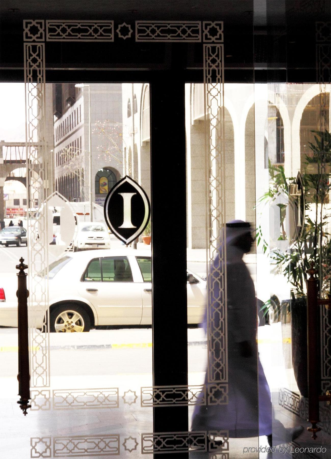 Intercontinental Dar Al Iman Madinah By Ihg Hotell Medina Inreriör bild The photo shows a view through a glass door or window, featuring an elegant design with a prominent "I" symbol displayed. Through the glass, there are some parked vehicles visible, including a white car. In the foreground, a shadowy figure of a perso