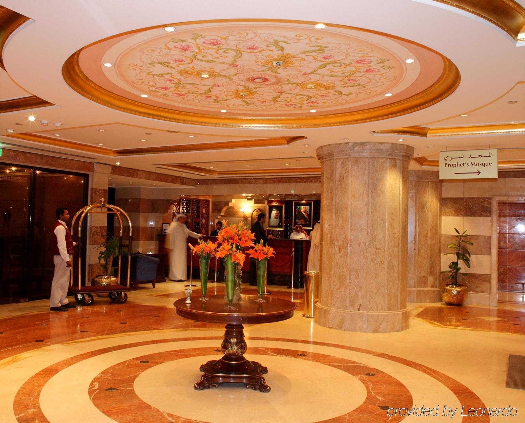Intercontinental Dar Al Iman Madinah By Ihg Hotell Medina Inreriör bild The photo depicts a luxurious hotel lobby. The ceiling features an ornate design with floral patterns, adding elegance to the space. In the center of the lobby, there is a decorative table with a bowl or vase of bright orange flowers. The flooring is
