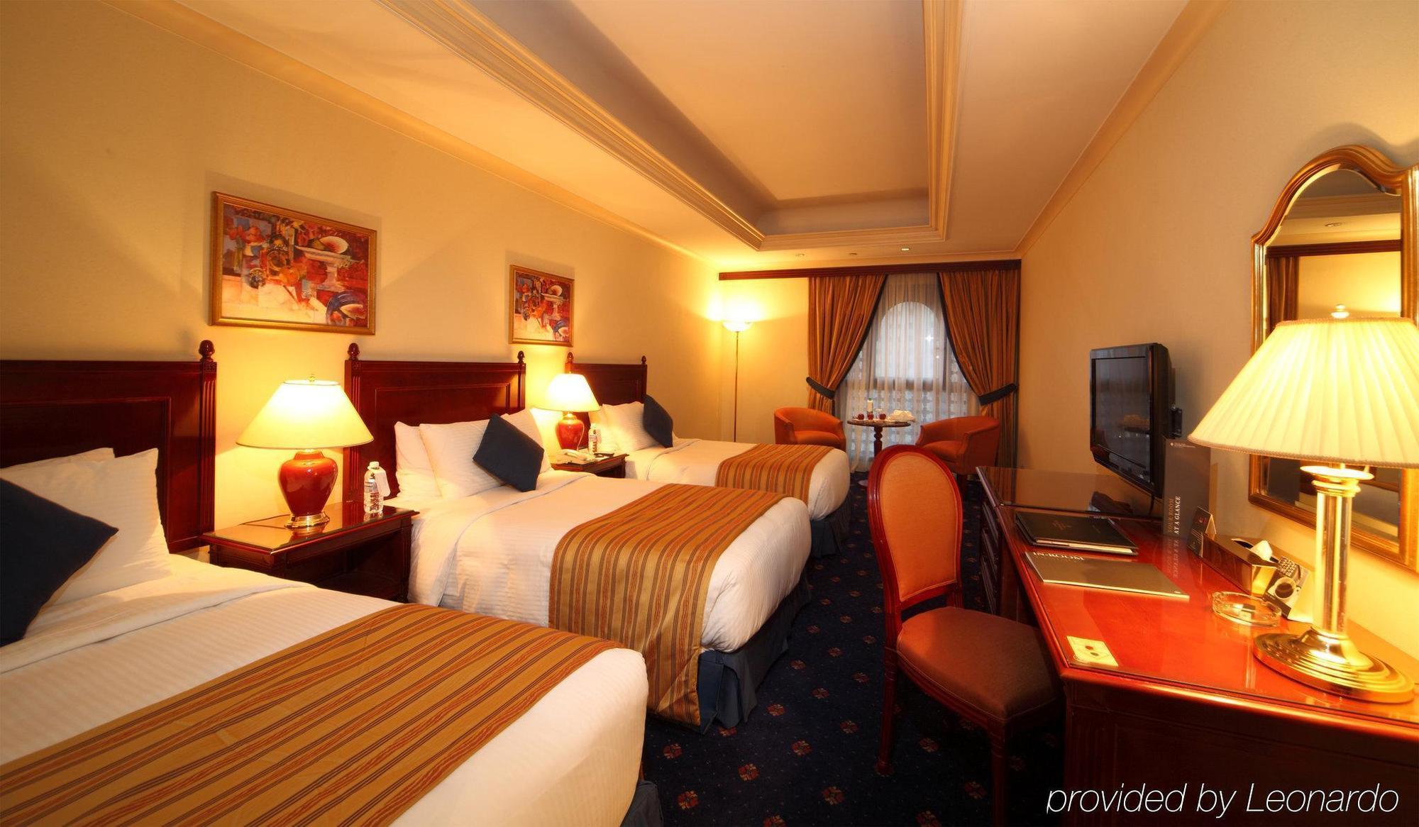 Intercontinental Dar Al Iman Madinah By Ihg Hotell Medina Rum bild The photo shows a hotel room with a warm and inviting atmosphere. There are two beds with white and gold bedding, complemented by decorative pillows. Each bed has a bedside lamp. In the background, there's a small seating area with a couple of chairs