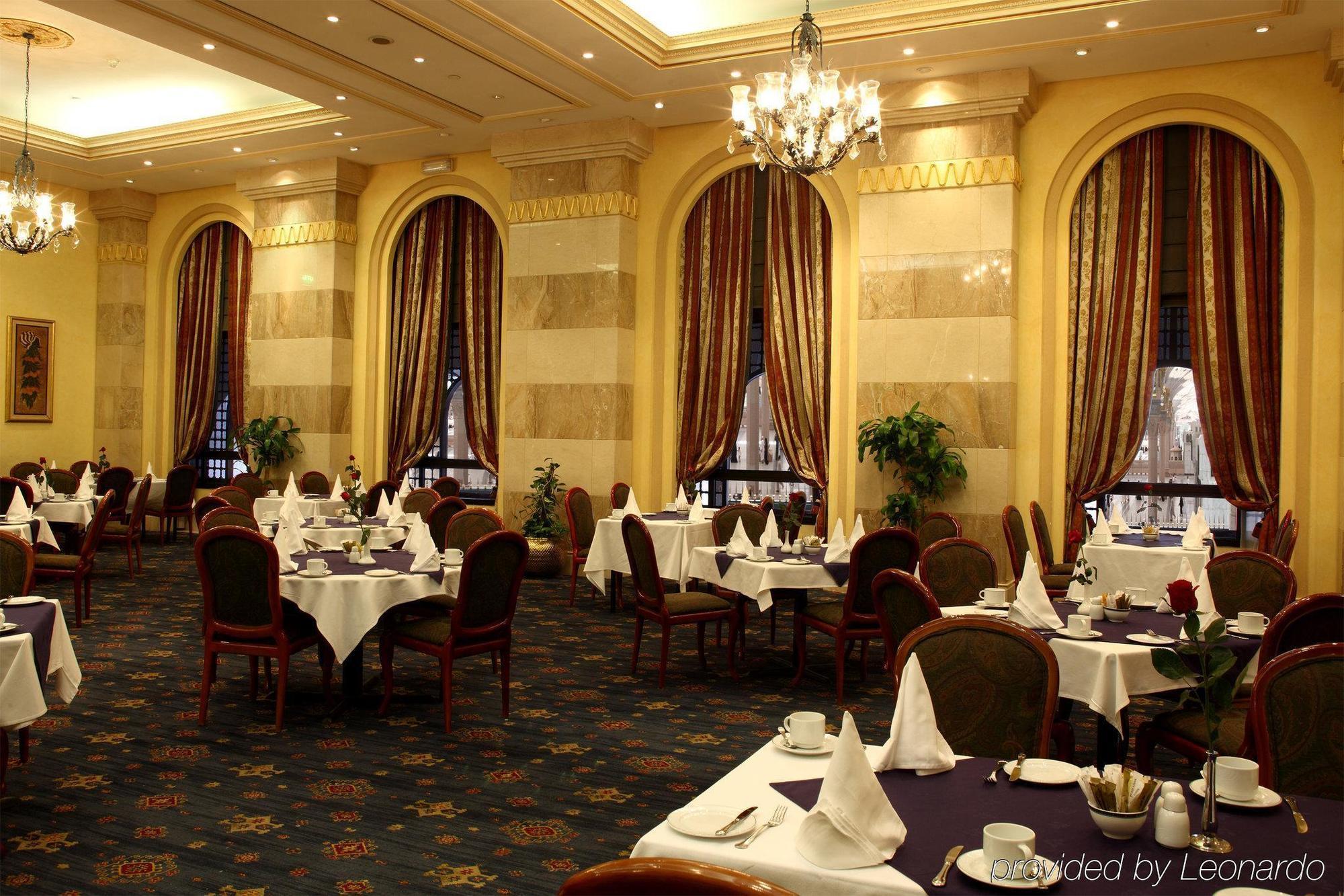 Intercontinental Dar Al Iman Madinah By Ihg Hotell Medina Restaurang bild The photo depicts an elegant restaurant interior. The space features ornate decor with tall windows draped in rich, patterned curtains. Tables are neatly set with white tablecloths and napkins, and there are plush chairs arranged around them. The flo