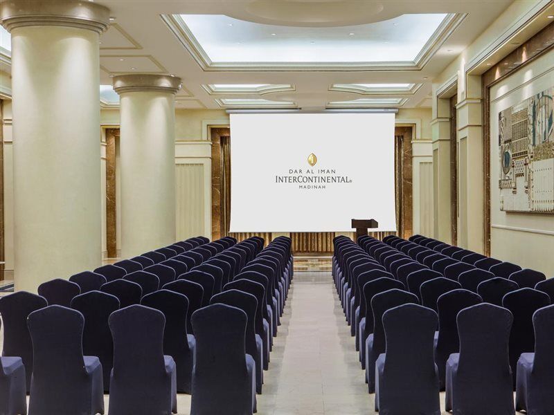 Intercontinental Dar Al Iman Madinah By Ihg Hotell Medina Exteriör bild The photo shows a conference or meeting room set up. There are rows of dark blue chairs arranged facing a large screen at the front of the room. The screen displays the logo of the Intercontinental hotel, indicating that this space is likely intended