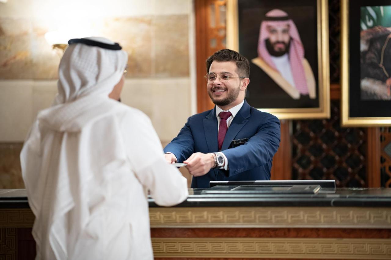 Intercontinental Dar Al Iman Madinah By Ihg Hotell Medina Exteriör bild The photo shows a hotel reception area. A male receptionist, dressed in a navy suit with a tie, is smiling and handing an item to a guest who is wearing traditional white attire, possibly a kandura or thobe. The background features elegantly decorate