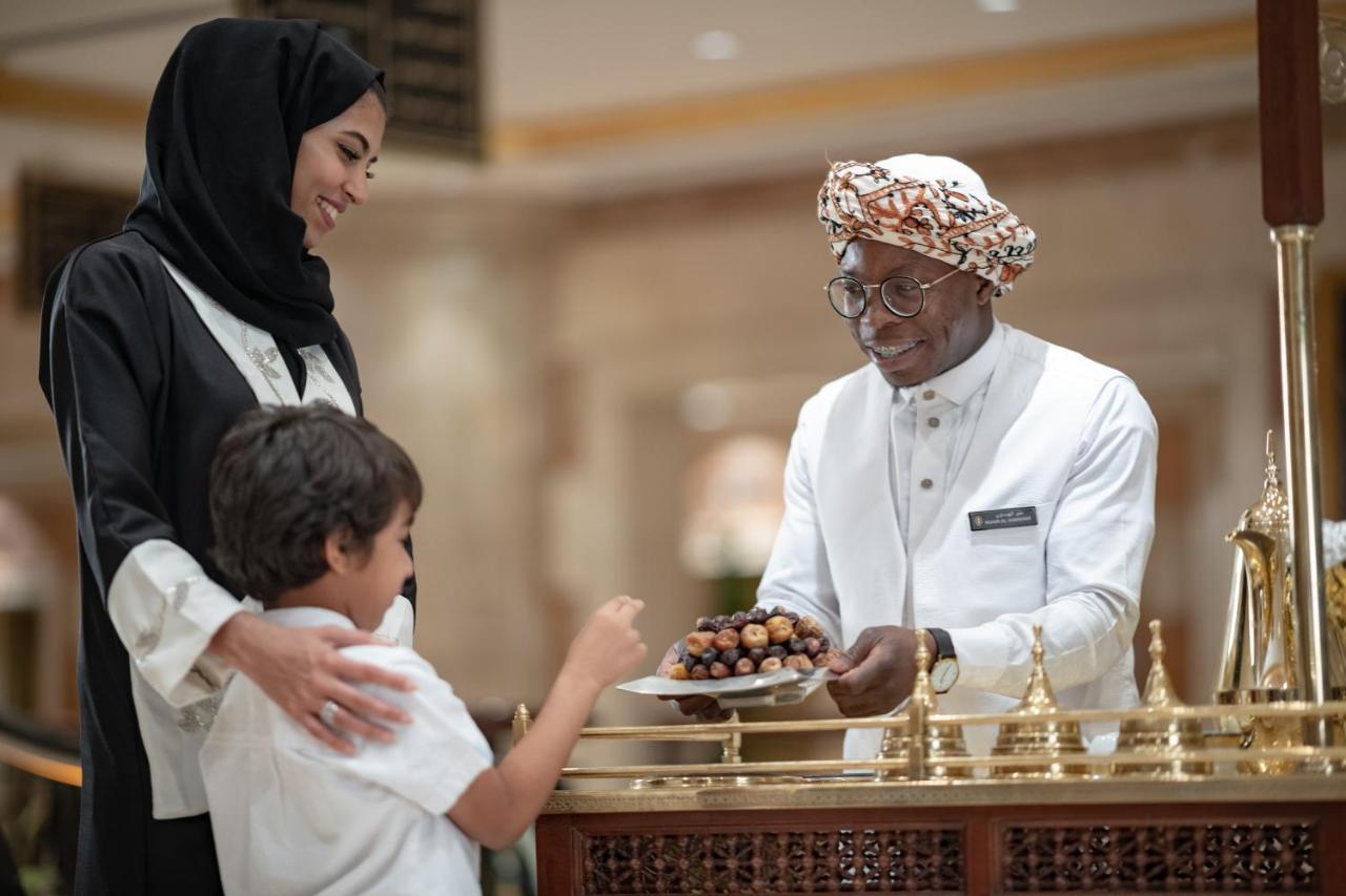 Intercontinental Dar Al Iman Madinah By Ihg Hotell Medina Exteriör bild The photo depicts a warm interaction between three people. A woman, dressed in a traditional black attire and a headscarf, stands beside a young boy who is reaching out towards a plate of dates held by a smiling man in a white traditional outfit with